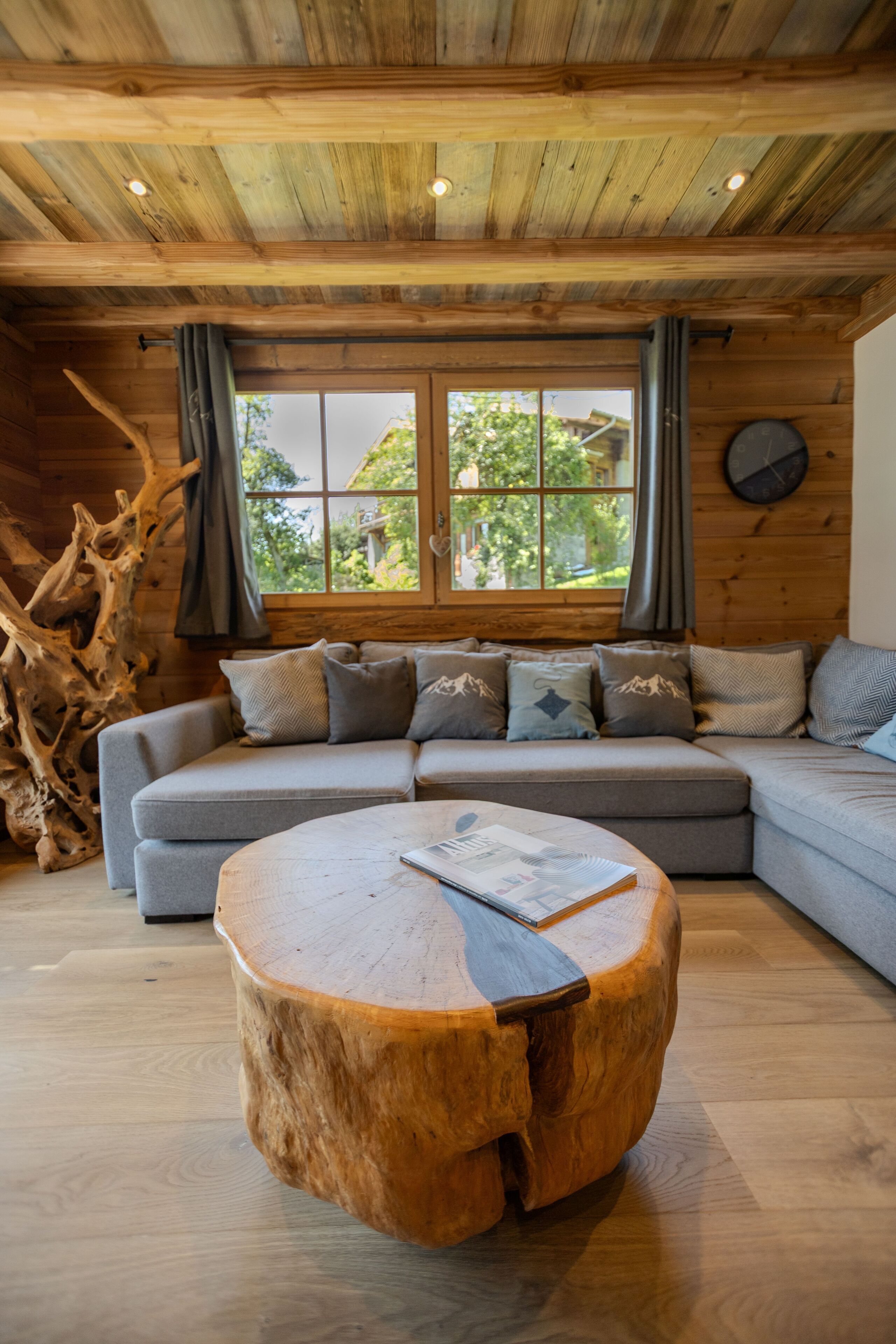 Una sala de estar acogedora con techo de madera, un ventanal con cortinas, un sofá seccional gris con cojines estampados y una mesa de centro redonda de madera.