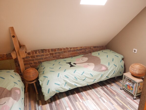Un dormitorio en ático acogedor con techo inclinado, paredes de ladrillo visto y una escalera de madera. Hay dos camas con ropa de cama verde a juego y una pequeña cesta de mimbre en el suelo.