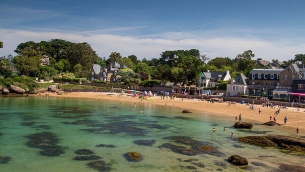 Uma vila costeira com uma praia de areia, águas claras com rochas e edifícios ao longo da costa.