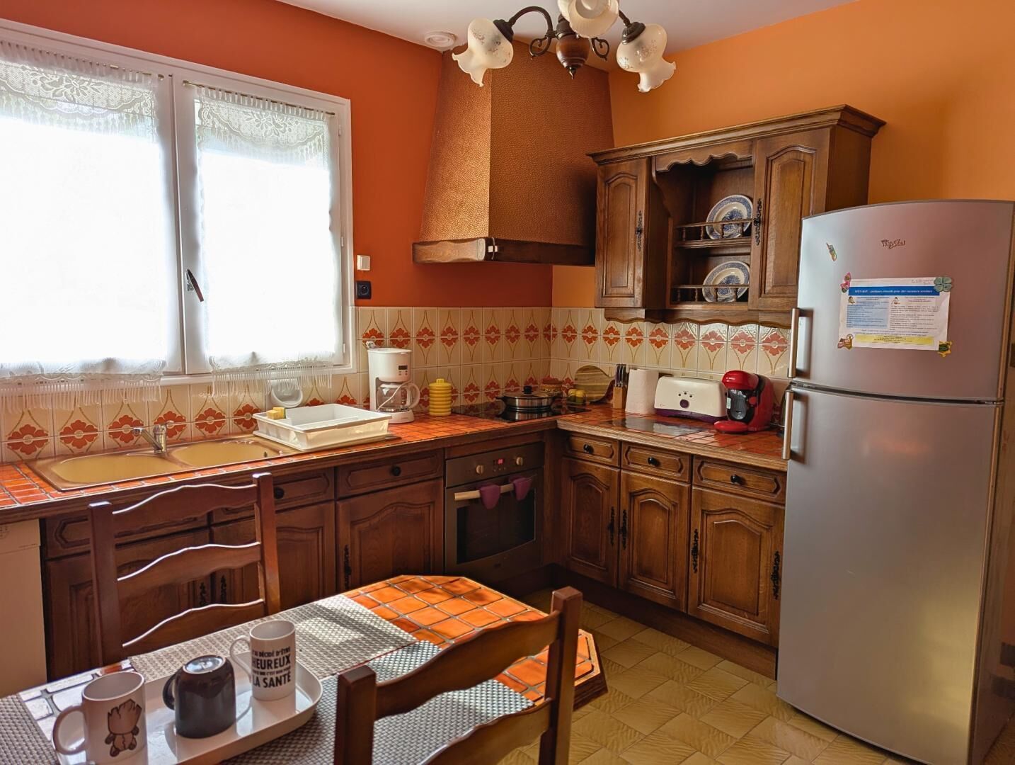 Une cuisine avec des meubles de rangement en bois, un réfrigérateur, une machine à café et une fenêtre ornée de rideaux en dentelle.