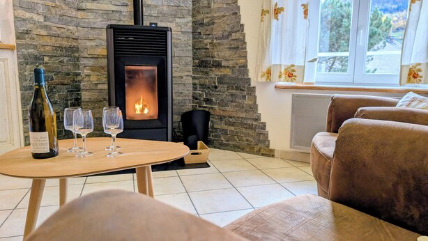 A cozy living room with a stone fireplace, a wooden table with a bottle of wine and glasses, and a view of the outdoors through a window. - Renovated stone house, classified 3 stars between La Bresse and Gérardmer (Basse-Sur-Le-Rupt)