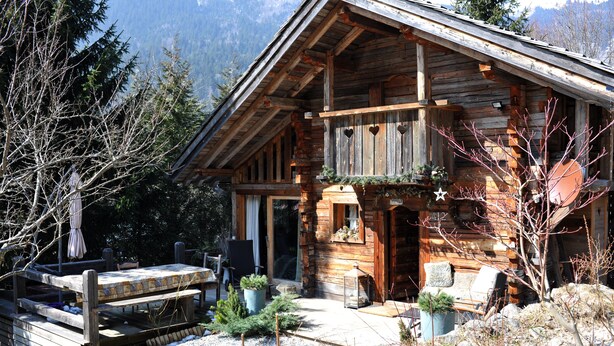 Un chalet rustique en rondins de bois, doté d’un balcon, d’une terrasse en bois et d’un chemin de pierre menant à l’entrée.