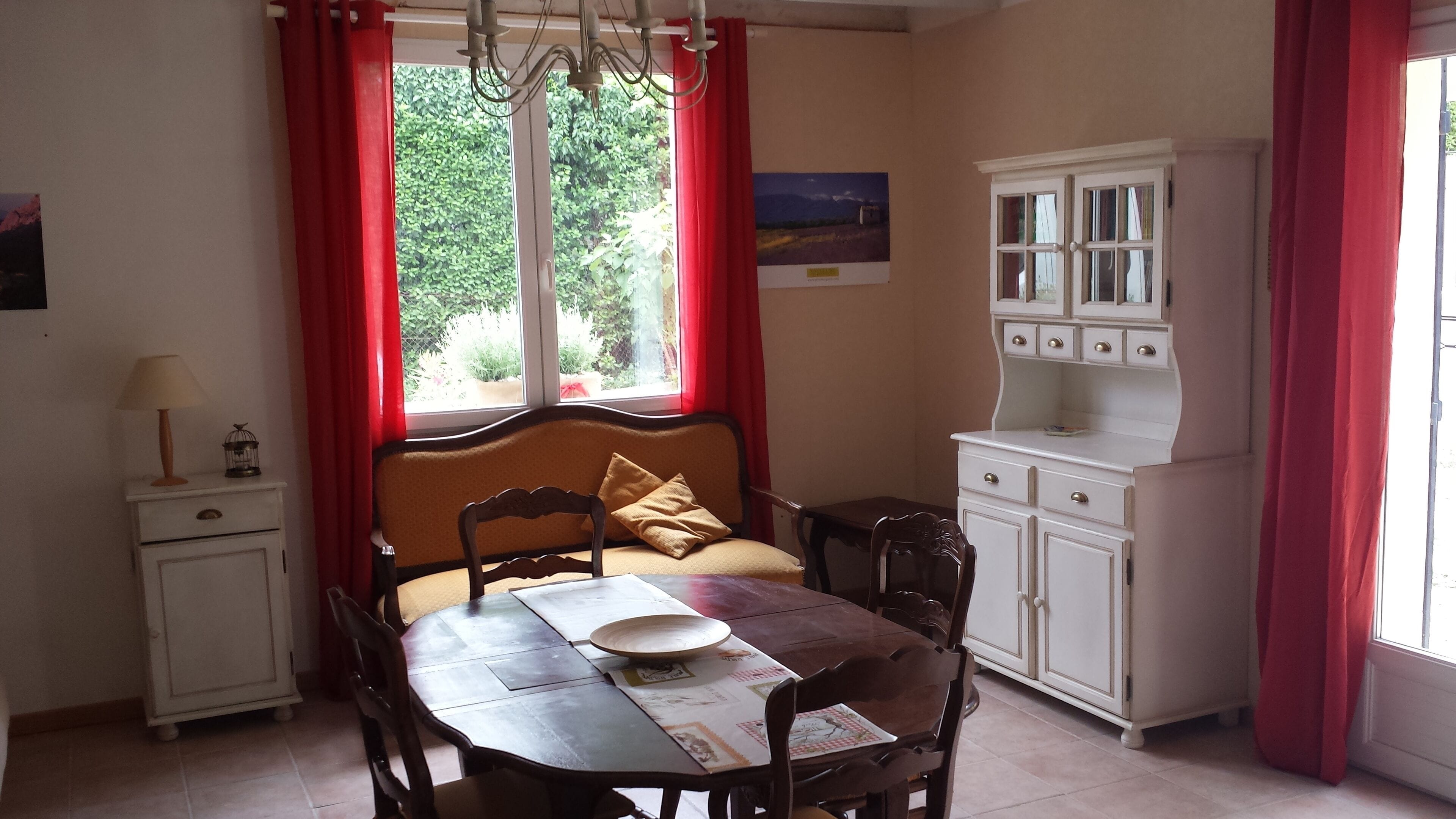 Un coin repas avec une table en bois, des chaises, une fenêtre avec des rideaux rouges, un meuble blanc et une petite table d’appoint avec une lampe.