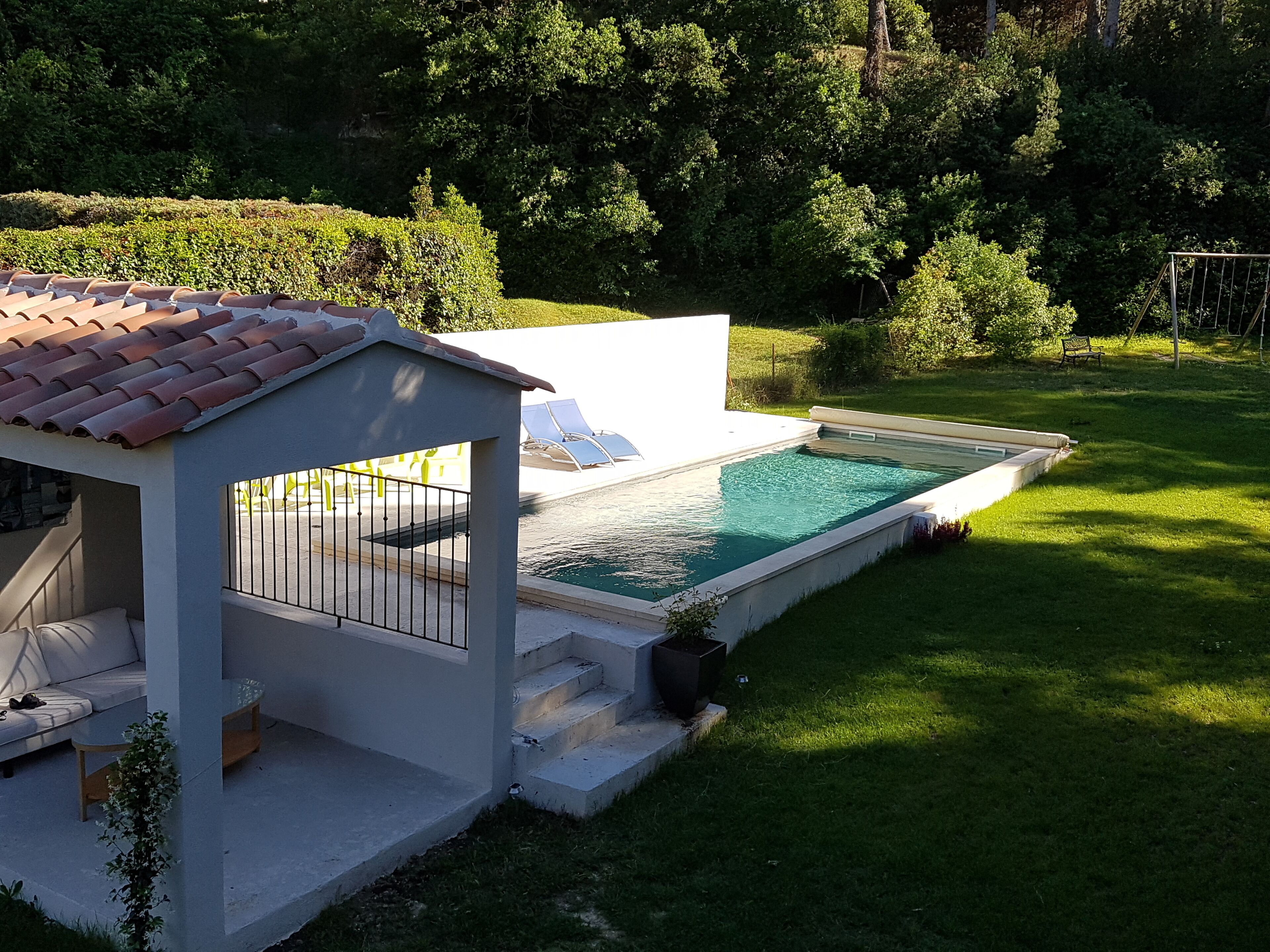 Un espace piscine avec un petit bâtiment blanc, un coin salon abrité et une pelouse verdoyante.