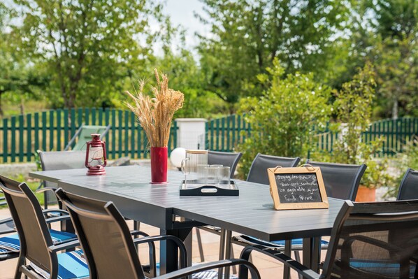 An outdoor dining area with a table set for two, surrounded by chairs. A red lantern and a vase with dried plants are on the table. A chalkboard menu is visible.