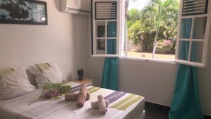 A neatly made bed with pillows, a teddy bear, and a bouquet of flowers. There is a window with curtains, an air conditioning unit, and a framed picture on the wall. - VILLA WITH PRIVATE POOL & GARDEN (8 PEOPLE) IN ST FRANCOIS GUADELOUPE (saint françois)