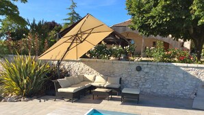 Zona junto a la piscina con un muro de piedra, un muelle de madera y una sombrilla color beige que da sombra.