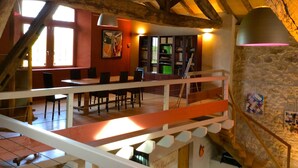 Un interior rústico con vigas de madera a la vista, una zona de comedor con mesas y sillas, y una escalera que conduce a un nivel superior.