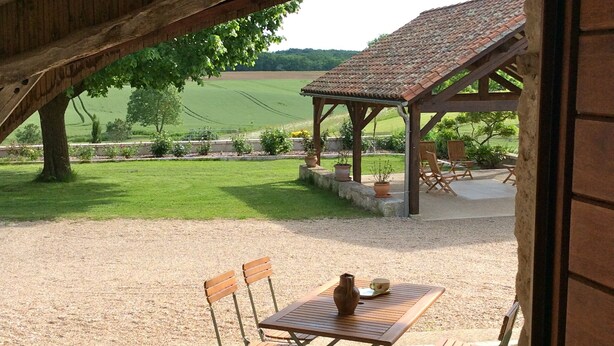 Un área de estar al aire libre con una mesa y sillas bajo una estructura de madera, con vista a un campo cubierto de césped y un edificio con techo de tejas.