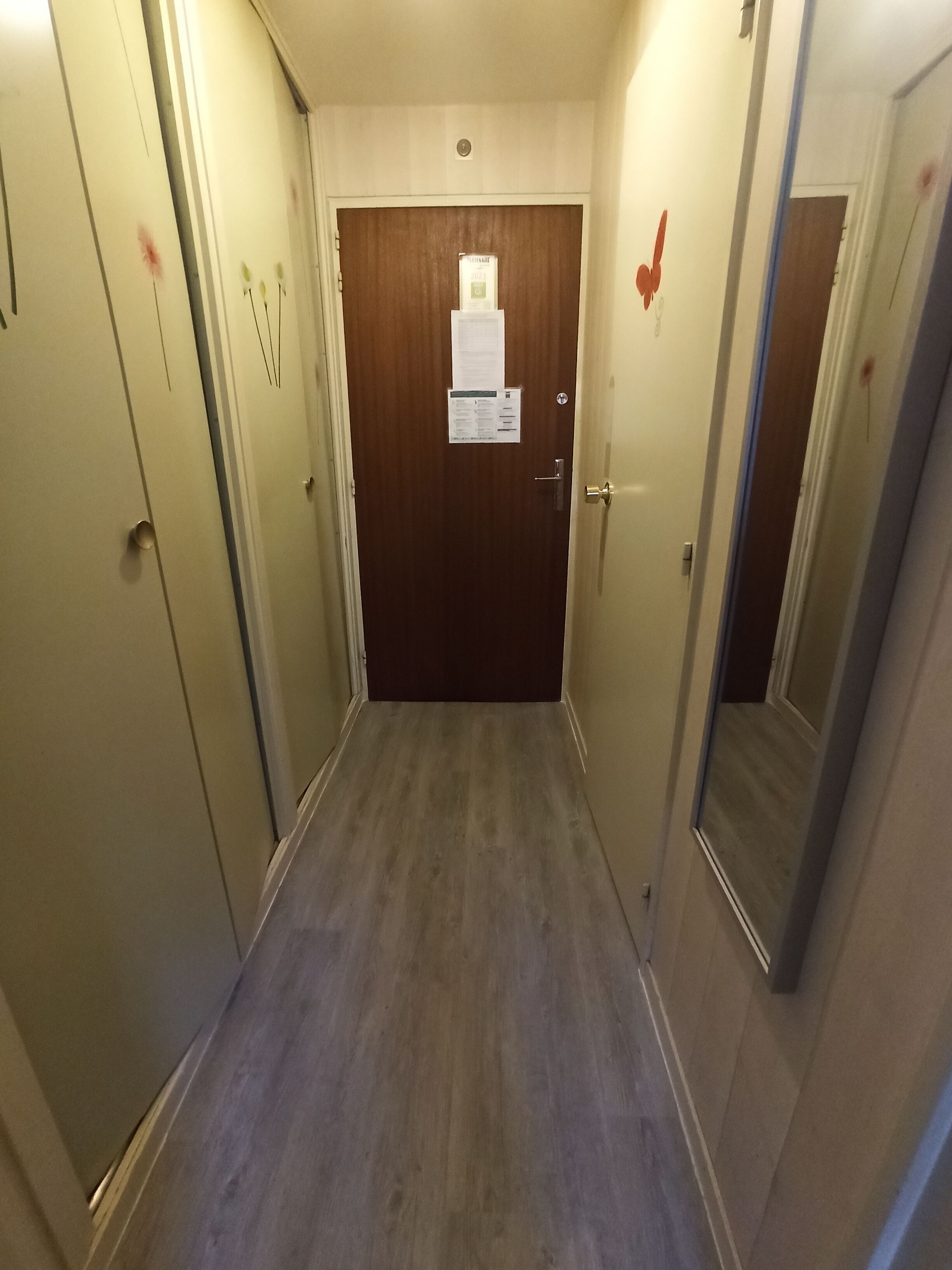 Een smalle gang met houten deuren aan weerszijden, een prikbord aan de rechterwand en een houten vloer.