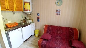 Une cuisine de taille réduite, équipée d’un évier, d’un petit réfrigérateur et de placards en bois. Un canapé rouge avec des coussins est placé contre le mur, et une poubelle jaune est visible.