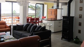 Ein modernes Wohnzimmer mit einer schwarzen Couch, einem Glastisch und einem Essbereich mit roten Stühlen und einem Holztisch. Eine Küche mit einem schwarzen Herd und weißen Schränken ist ebenfalls zu sehen.