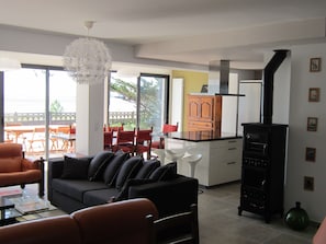 Un salon moderne avec un canapé noir, une table basse en verre et un coin repas avec des chaises rouges et une table en bois. On peut apercevoir une cuisine avec un four noir et des placards blancs.