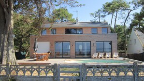 Ein zweistöckiges Haus mit einem großen Glasfenster, einem Swimmingpool und einer Holzterrasse.