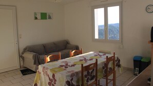 A dining area with a table covered in a floral tablecloth, surrounded by wooden chairs, a sofa, and a window with a view of the outdoors. - Furnished tourist accommodation 3 * south ardèche with shared pool (Rosières)