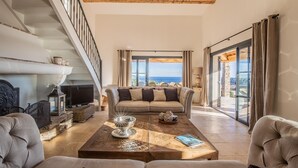 A spacious living room with a high ceiling, a staircase, a large window with a view, and a cozy seating area. - VILLA SOLEIL D'OR GOLFE DE ST TROPEZ4*, swimming pool at 29° all year round, 180° view (Cavalaire-sur-Mer)