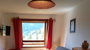 Una habitación acogedora con un sofá de madera, una mesita pequeña, una silla roja y un armario de madera. Una ventana con cortinas rojas ofrece vistas a un paisaje montañoso.