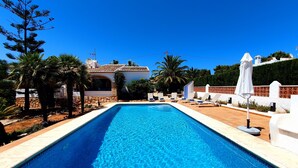 Une piscine rectangulaire à l’eau d’un bleu limpide, entourée d’une terrasse carrelée. On y trouve des chaises longues et des parasols sur le côté droit, ainsi que des palmiers et un mur de pierre sur le côté gauche.