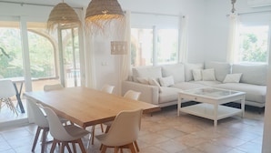 Un salon spacieux avec une table à manger en bois, des chaises blanches, un canapé blanc et une table basse. De grandes fenêtres laissent entrer la lumière naturelle et offrent une vue sur la verdure environnante.