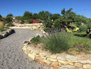 Ein Garten mit Kiesweg, Steinmauern und verschiedenen Pflanzen, darunter Lavendel. Im Hintergrund stehen gelbe Stühle und ein Tisch.