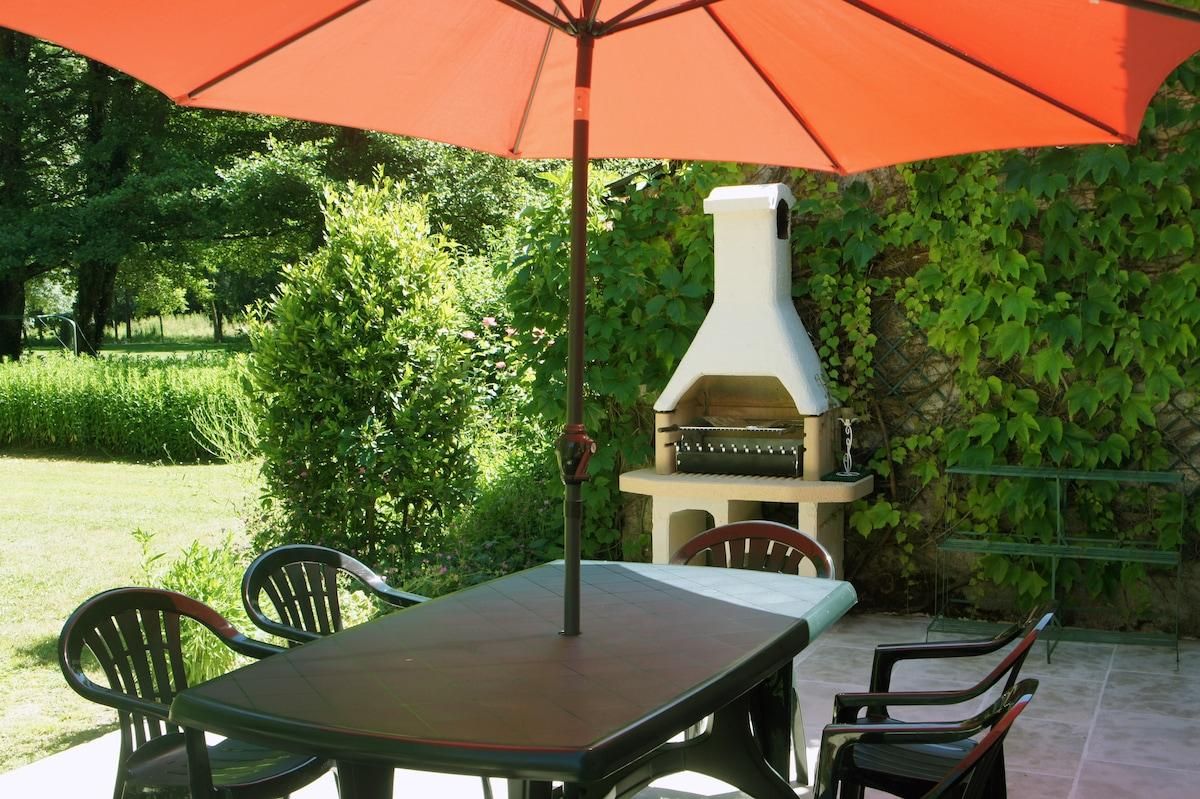 Een terras buiten met een barbecue, een tafel en stoelen onder een rode parasol.