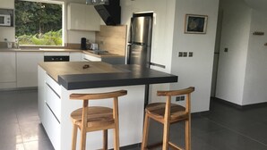 Una cucina moderna con isola centrale, due sgabelli in legno, elettrodomestici in acciaio inossidabile e vista su spazi verdi.