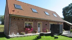 A two-story brick house with a sloped roof, two skylights, a front door, and a small porch area with potted plants and a chair. - Discover our farm cottage, visit livestock (Rodelinghem)