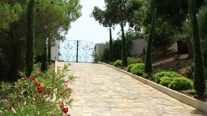 Un chemin de pierre mène à une entrée fermée par un portail, qui est elle-même entourée de verdure et de plantes en fleurs.