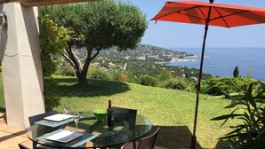 Une table en verre, des chaises et un parasol rouge, le tout offrant une vue sur un paysage côtier, avec la mer et une ville au loin.