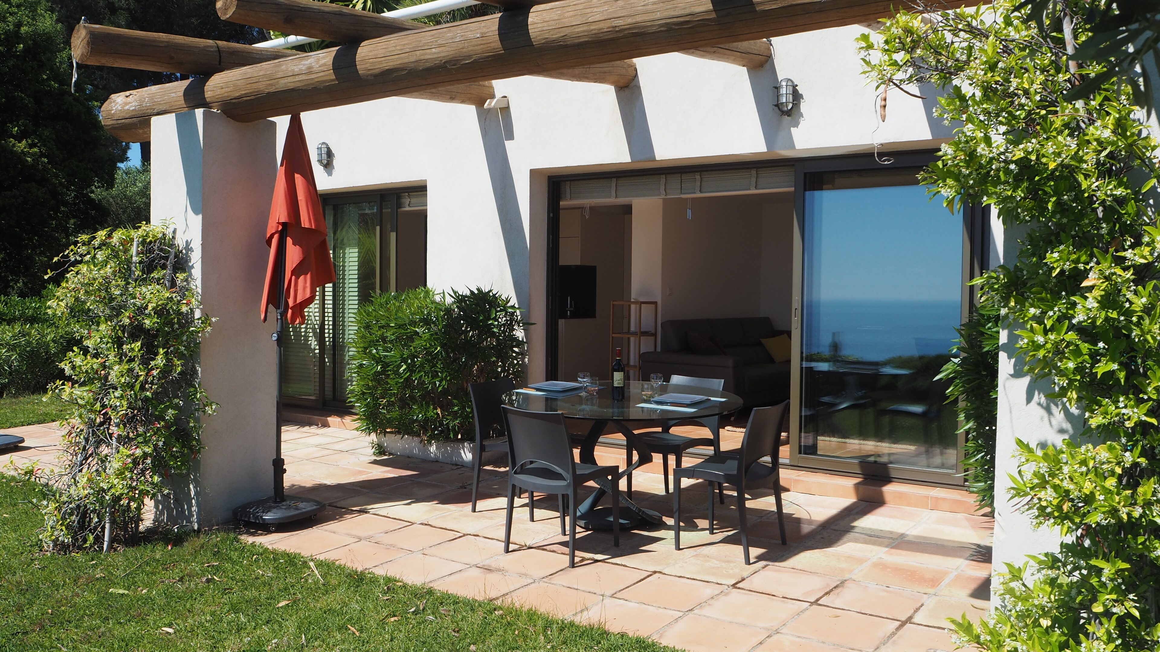 A patio with a table and chairs, a red umbrella, and a view of the sea through a glass door.