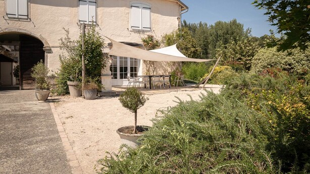 Una casa grande con un porche cubierto, plantas en macetas y un árbol pequeño.
