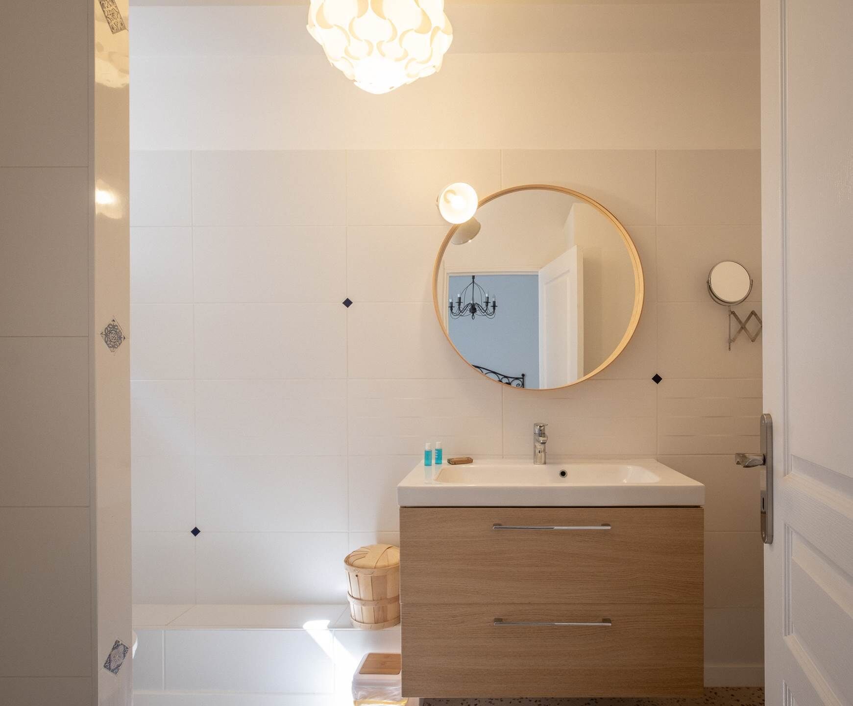 A modern bathroom with a round mirror, white tiles, a wooden vanity, and a light fixture.
