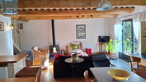 A cozy living room with a wooden ceiling, a fireplace, wicker furniture, a dining table, and a large window with curtains.
