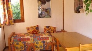 A small room with a bed, a wooden table, and a chair. There is a window with curtains, a wall-mounted picture, and a plant on the wall.