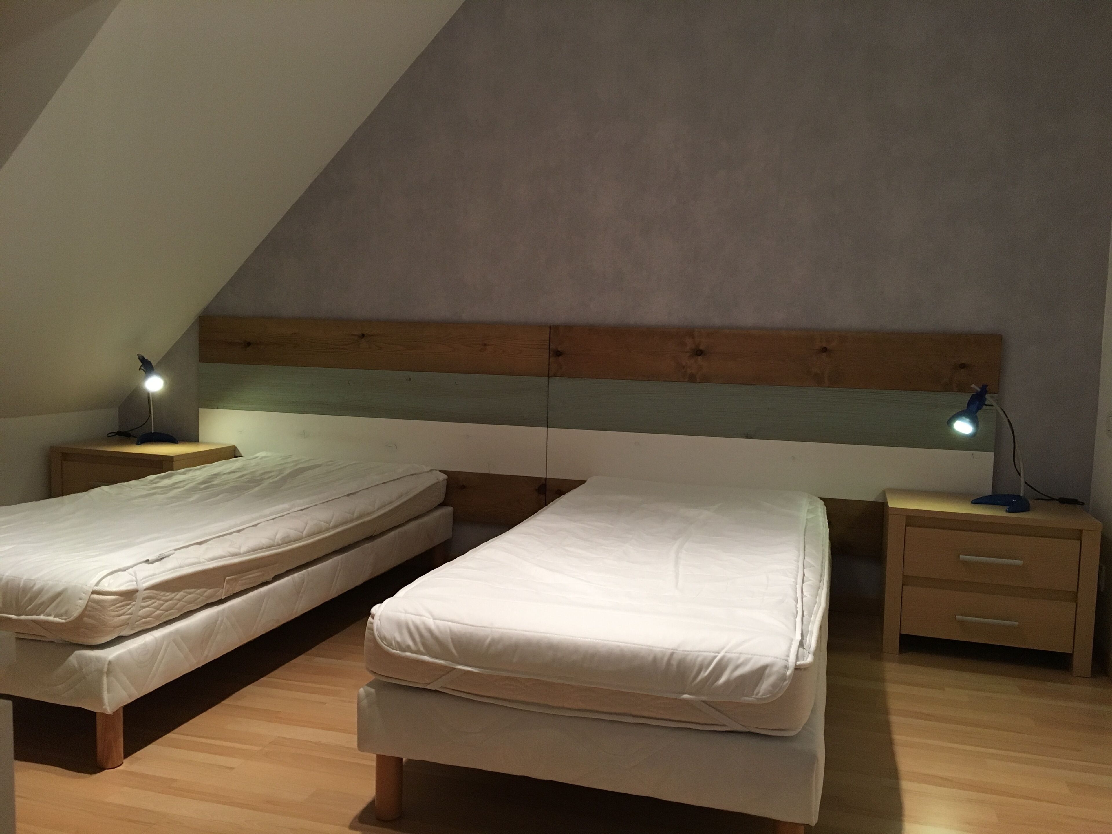 A room with two single beds, wooden headboards, bedside tables with lamps, and a wooden floor.