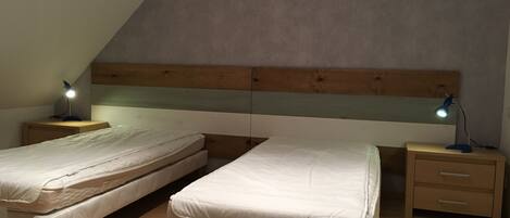 A room with two single beds, wooden headboards, bedside tables with lamps, and a wooden floor.