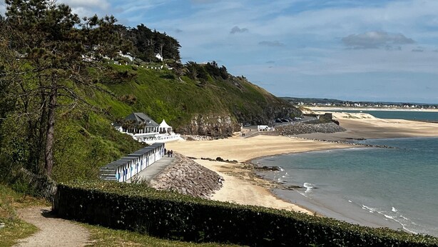 Un paisaje costero con una playa, un edificio blanco con techo azul y una colina con vegetación y casas.