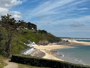 A coastal landscape with a beach, a white building with a blue roof, and a hill with greenery and houses.