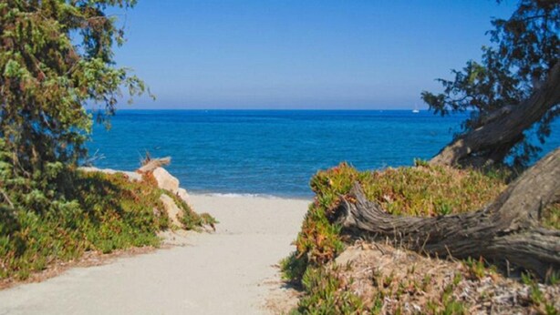 Camino costero que lleva a una playa con agua azul cristalina y un velero a lo lejos.