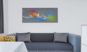 A modern living room with a grey sofa, two grey cushions, a colorful abstract painting on the wall, and a patterned yellow chair.