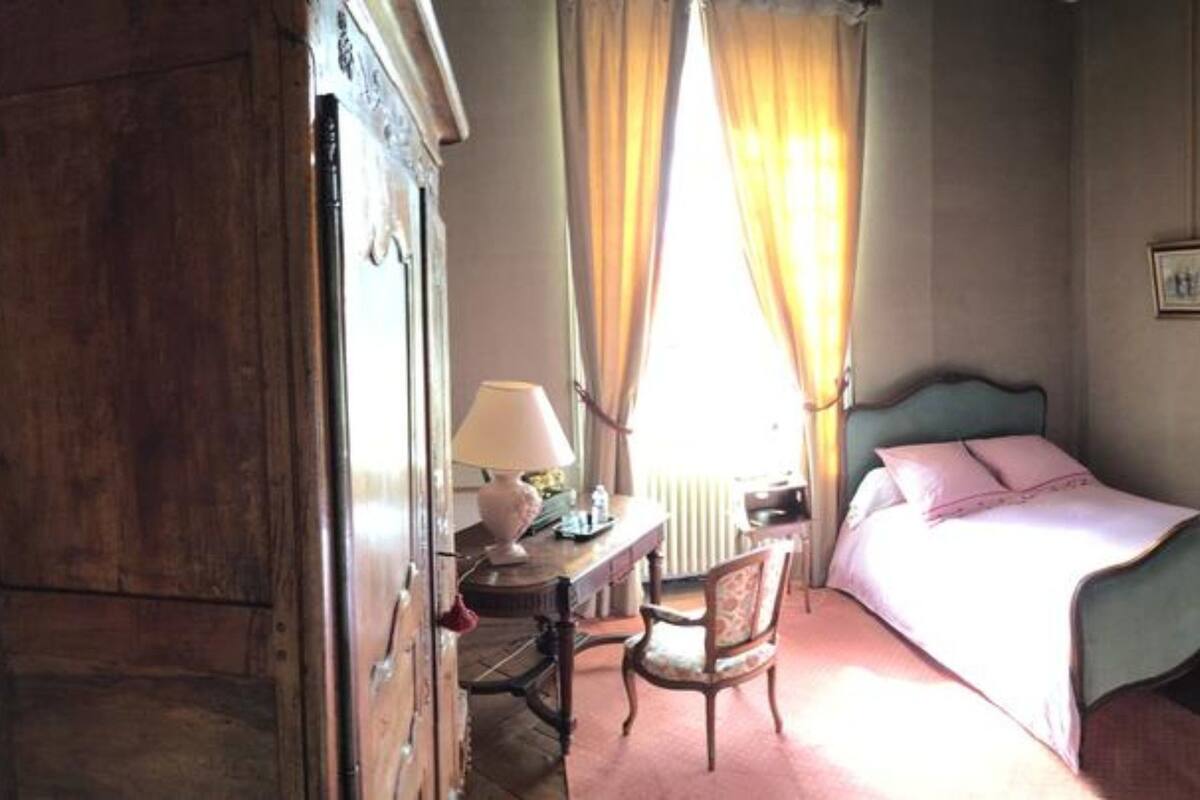 A bedroom with a bed, a chair, a desk with a lamp, a wardrobe, and a door leading to another room.