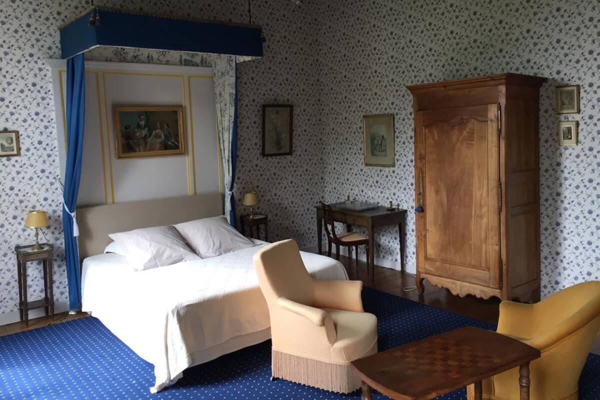 A bedroom with a four-poster bed, a wooden cabinet, a chair, a small table, and a blue patterned rug.