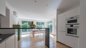 A modern kitchen with white cabinets, a dining area with a table and chairs, and a large window with a view of greenery. - Villa Olivier with a private pool—10 minutes from the beach (Bompas)