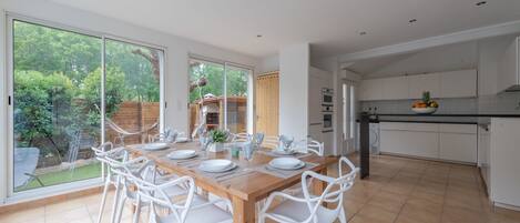 A spacious dining area with a wooden table and white chairs, large sliding glass doors leading to a garden, and a modern kitchen with white cabinets.