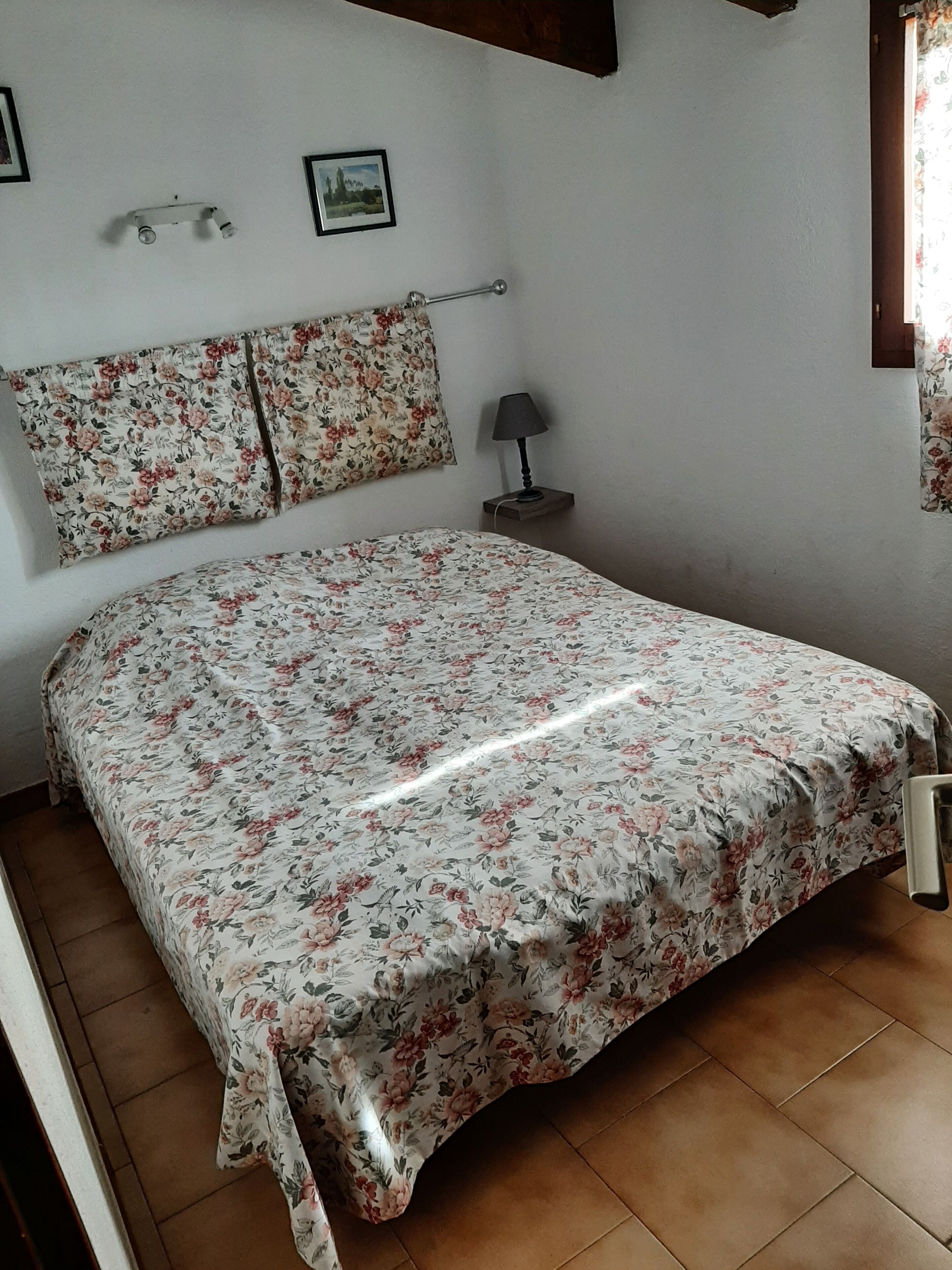 A bedroom with a floral bedspread, a headboard, a nightstand with a lamp, and a window with floral curtains.