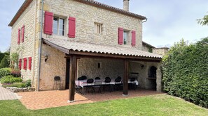 A stone building with red shutters, a covered patio with tables and chairs, and a brick oven. - House with private swimming pool, in the heart of south-west (Thézac)