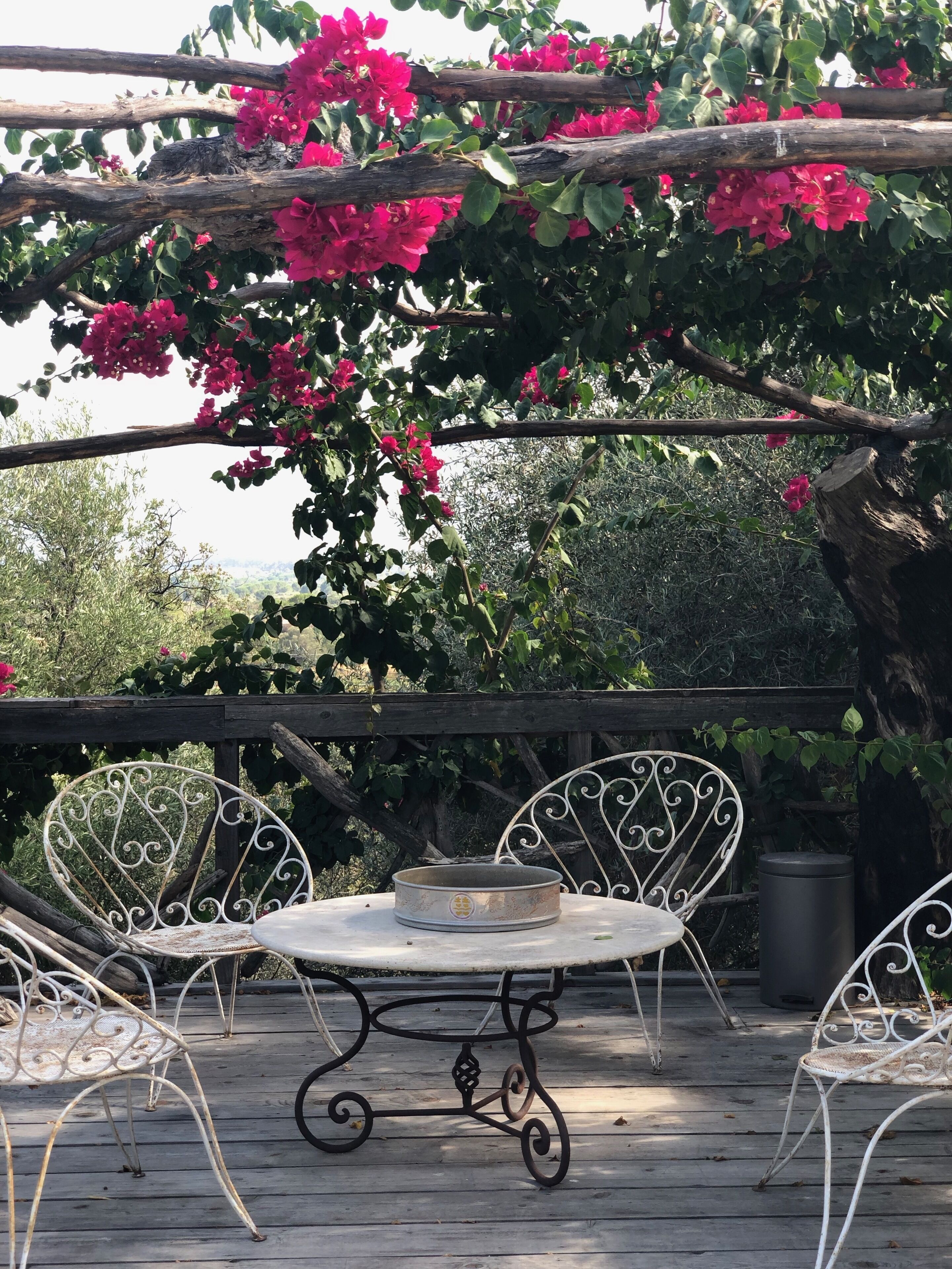 A wooden deck with a metal table and chairs, surrounded by lush greenery and pink flowers.