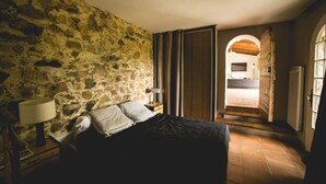 Une chambre avec un mur en pierre, un lit recouvert d’une couverture foncée, une table de chevet avec une lampe et une armoire en bois.