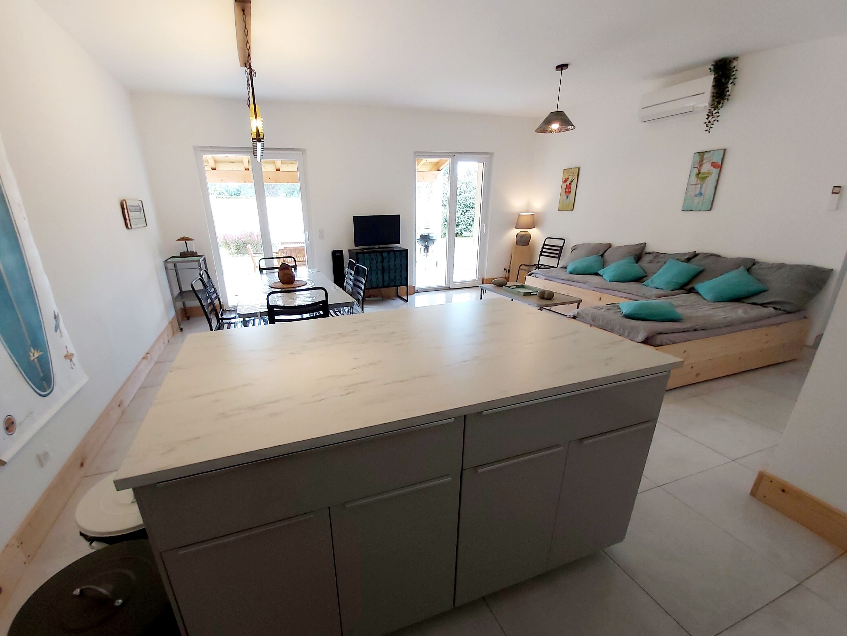 A modern kitchen with a central island, dining area with chairs, a sofa with teal cushions, and a TV.