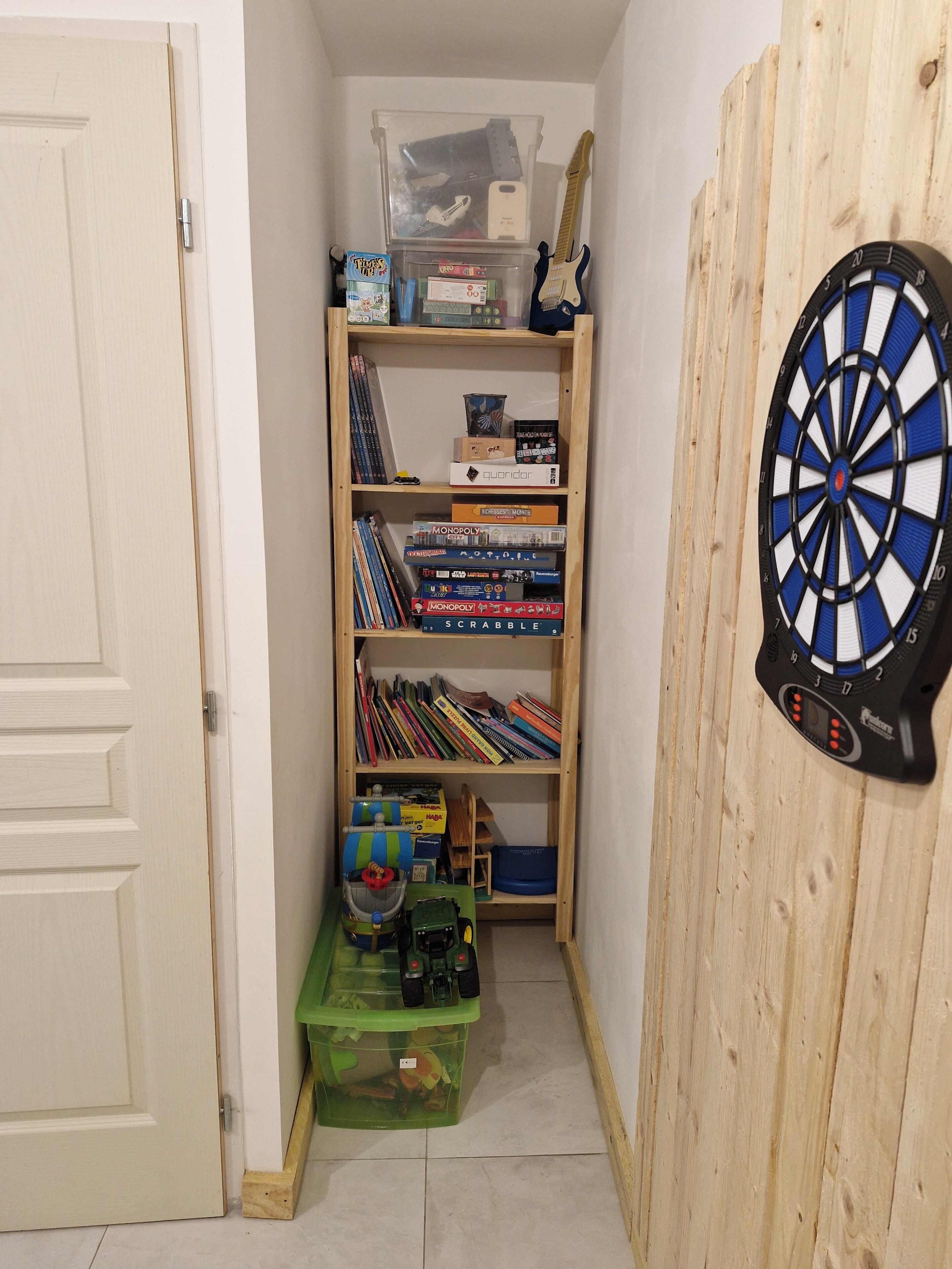 A room with a bookshelf containing board games, a dartboard, and a green storage bin.